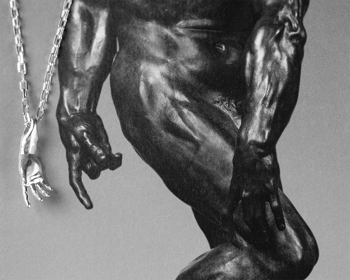 Sterling 925 silver pendant of Rodin's sculpture 'Hand of Adam' hanging from a chain, placed on a black-and-white background featuring a close-up of the same sculpture.