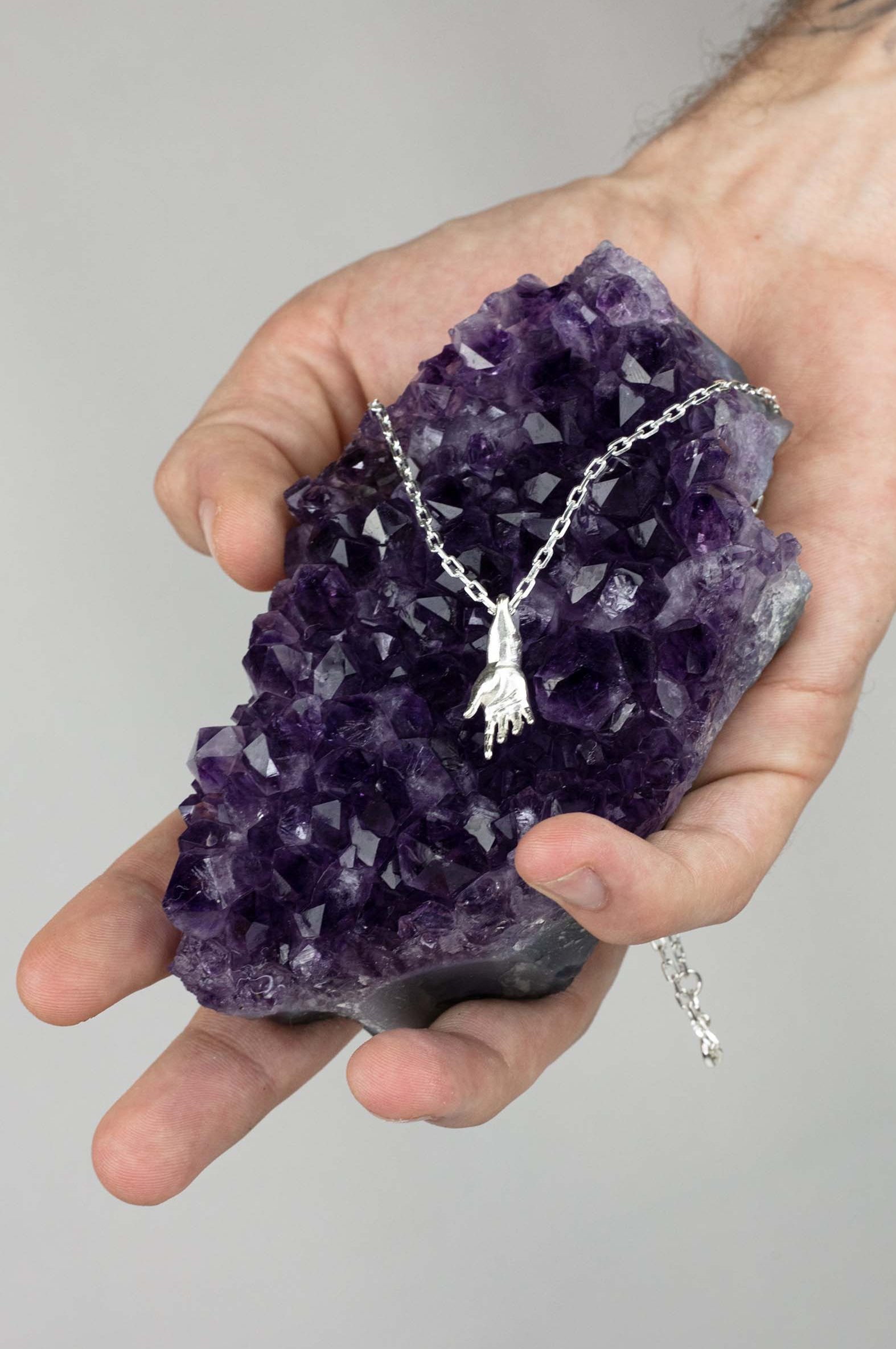 Sterling 925 silver pendant of Rodin's sculpture 'Hand of Adam' hanging from a chain, placed on a large amethyst crystal held in a hand.