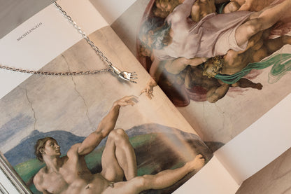 Sterling 925 silver pendant of Rodin's sculpture 'Hand of Adam' hanging from a chain, placed on an open book featuring a detail from Michelangelo's 'The Creation of Adam' fresco.