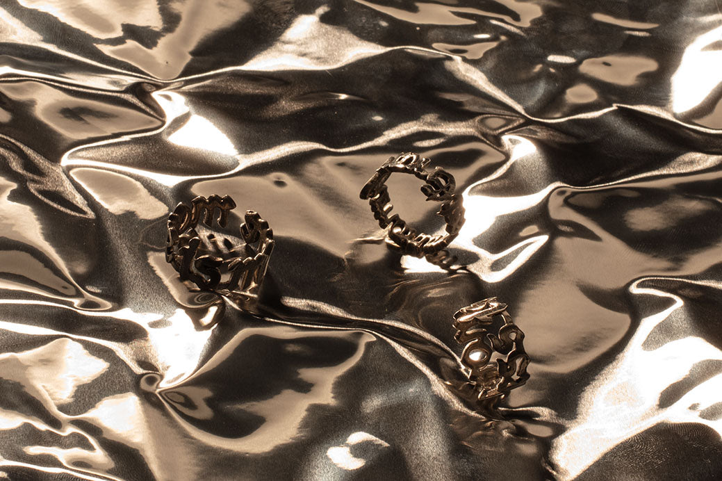 Two 'more is more' rings and one 'errare humanum est' ring, all made from Sterling 925 silver, arranged on a reflective, crumpled metallic background. The intricate shape on the rings stand out against the shiny, textured surface, highlighting their modern design.