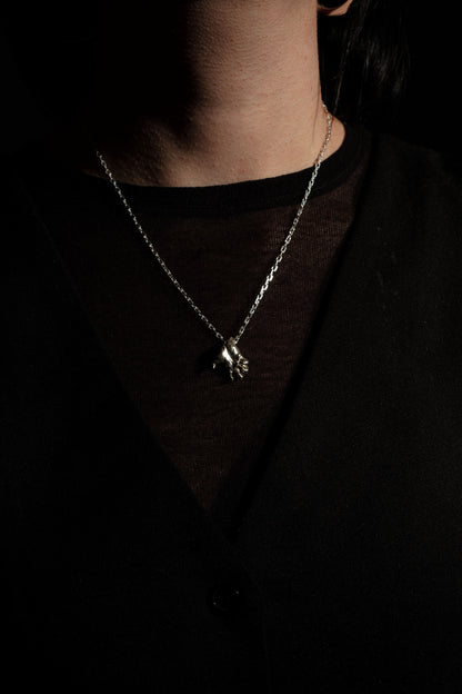 Sterling 925 silver pendant modeled after Rodin's sculpture 'Clenched Hand,' hanging from a delicate chain around the neck of the wearer. The pendant, depicting a clenched hand, is captured against a dark background, emphasizing its intricate design and craftsmanship.