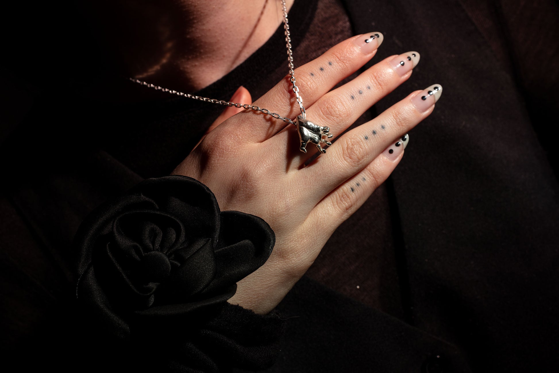 Sterling 925 silver pendant modeled after Rodin's sculpture 'Clenched Hand,' hanging from a chain around the neck of the wearer. One hand rests on their chest, adorned with tattoos and stylish black nails. A black rose accentuates the sleeve of the wearer's dark top, creating a bold contrast with the detailed pendant and lighting.