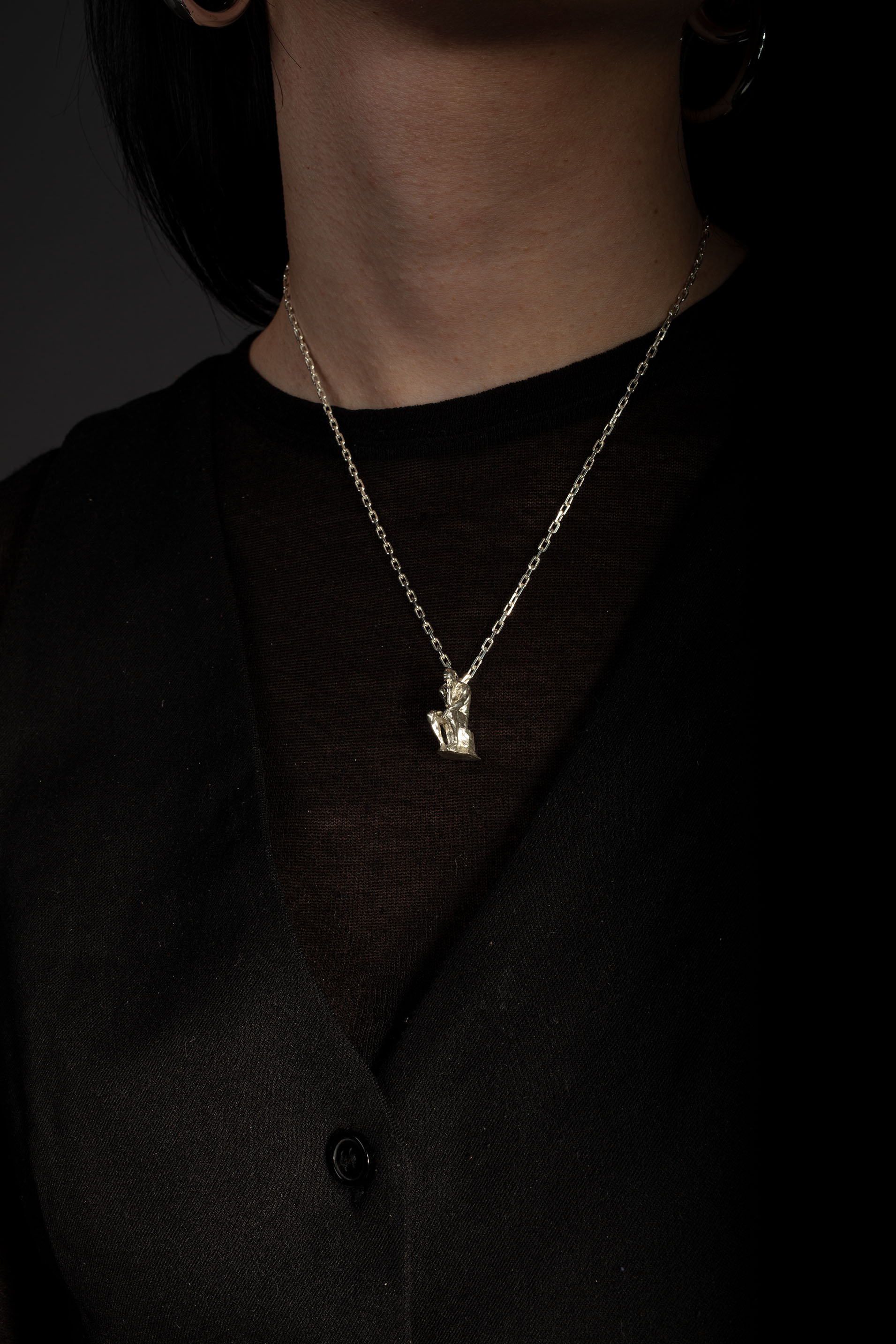 Sterling 925 silver pendant modeled after Rodin's sculpture 'The Thinker,' hanging from a delicate chain around the neck of the wearer, who is dressed in a black top. The pendant, depicting the seated figure in a contemplative pose, stands out against the dark background.