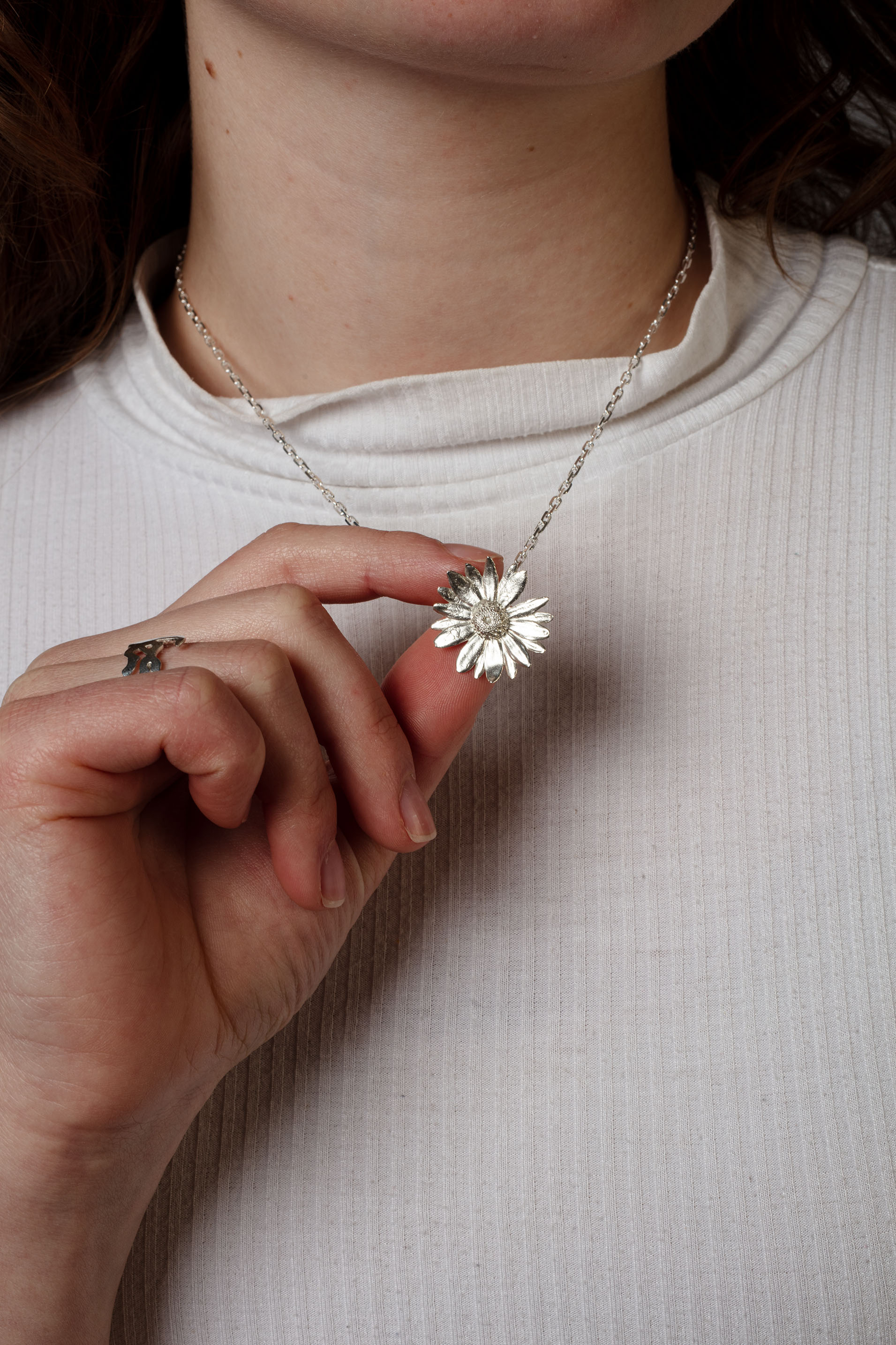 Sterling 925 silver pendant of a daisy flower, held in hand and hanging from a delicate silver chain. The pendant's intricate floral design is clearly visible, with the person wearing a white ribbed shirt, highlighting the contrast between the bright pendant and the soft texture of the fabric.