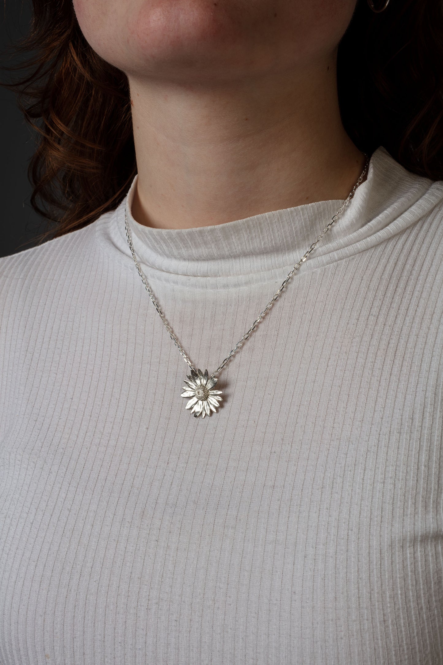 Sterling 925 silver pendant of a daisy flower, hanging from a delicate silver chain around the neck. The pendant's intricate floral design is clearly visible, with the person wearing a white ribbed shirt, highlighting the contrast between the bright pendant and the soft texture of the fabric.