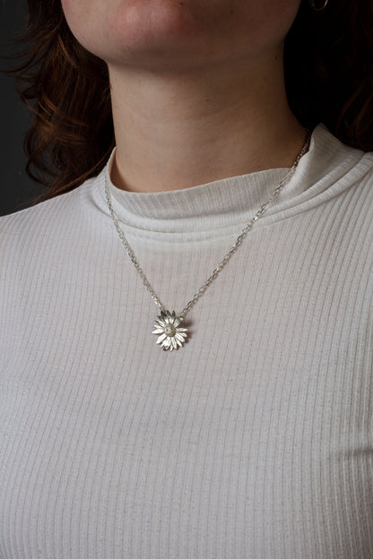 Sterling 925 silver pendant of a daisy flower, hanging from a delicate silver chain around the neck. The pendant's intricate floral design is clearly visible, with the person wearing a white ribbed shirt, highlighting the contrast between the bright pendant and the soft texture of the fabric.