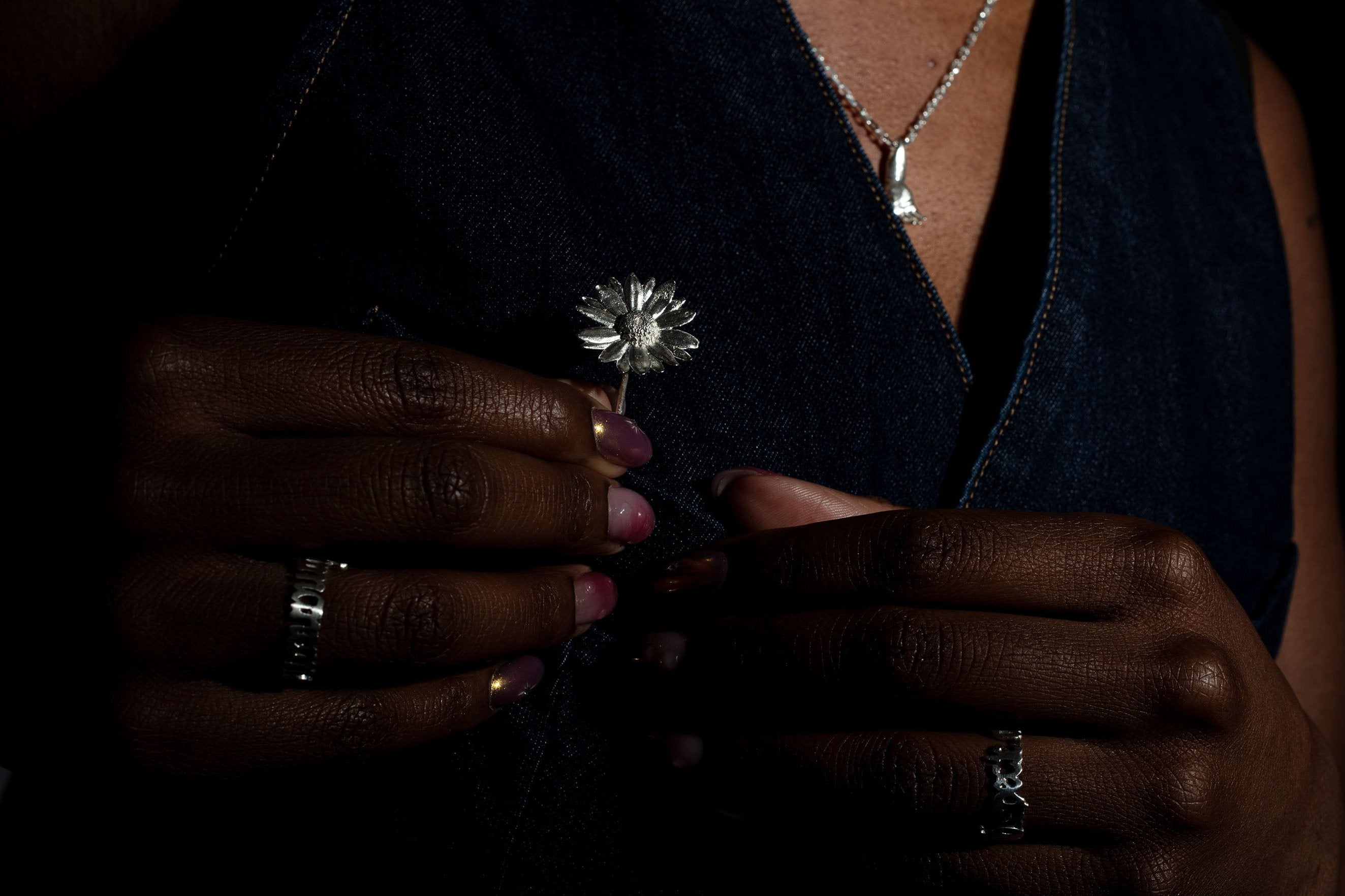 Person holding a Sterling 925 silver daisy brooch in one hand while wearing matching rings on their fingers, with a silver pendant necklace visible around their neck. The brooch is delicately held against a dark background, emphasizing its intricate floral design, while the rings add to the stylish composition.